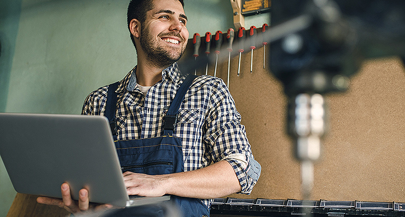Bildmotiv: Grinsender Mann mit Latzhose hält in einer Werkstatt einen Laptop in seiner Hand